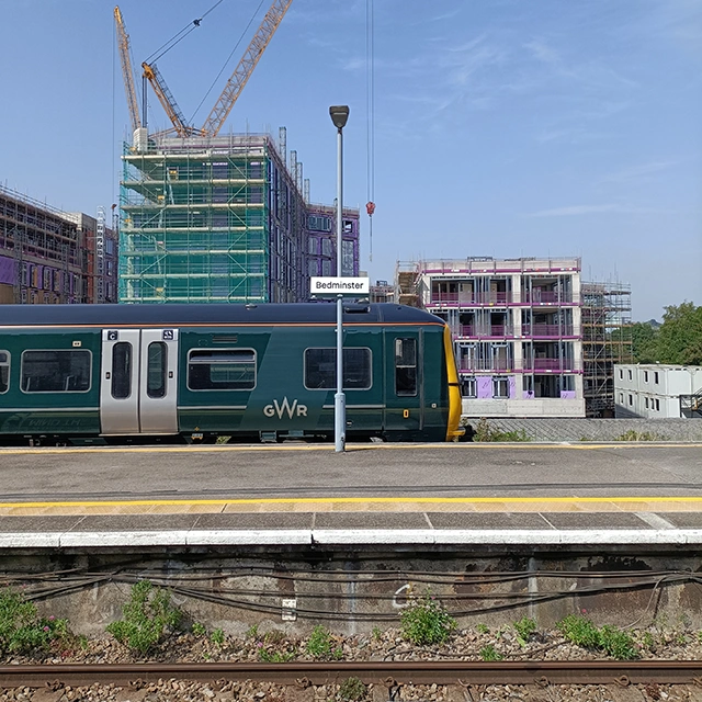 A green train arriving on the platform at Bedminster station. In the background is the Bedminster Green construction site.