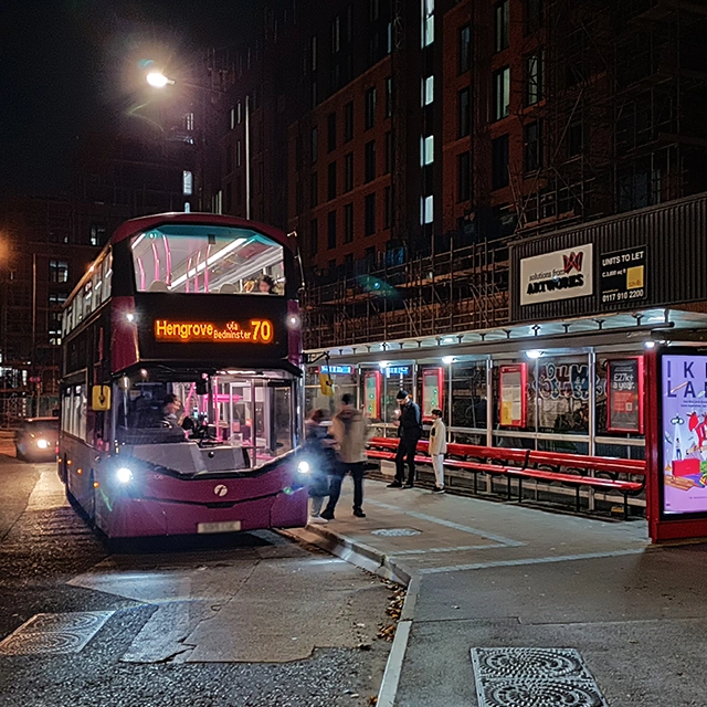 A pink double decker bus is pulled up at Dalby Avenue bus stop as two people wait to board.